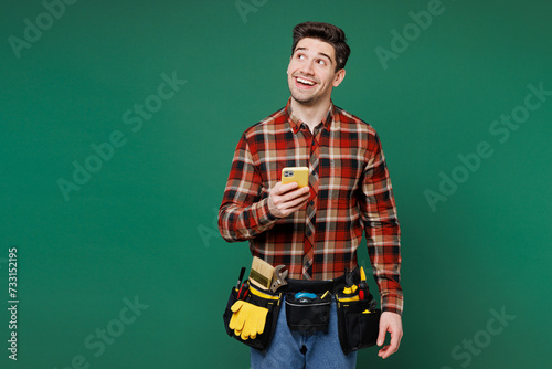 Young employee laborer handyman man wears red shirt hold use mobile cell phone look aside isolated on plain green background. Instruments accessories for renovation apartment room Repair home concept