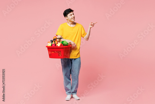 Young Caucasian man he wears yellow t-shirt casual clothes shopping hold in hand basket with food products point finger aside on area isolated on plain pastel light pink background. Lifestyle concept.