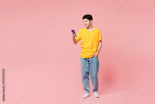 Full body smiling happy young man he wearing yellow t-shirt casual clothes hold in hand use mobile cell phone isolated on plain pastel light pink color background studio portrait. Lifestyle concept.