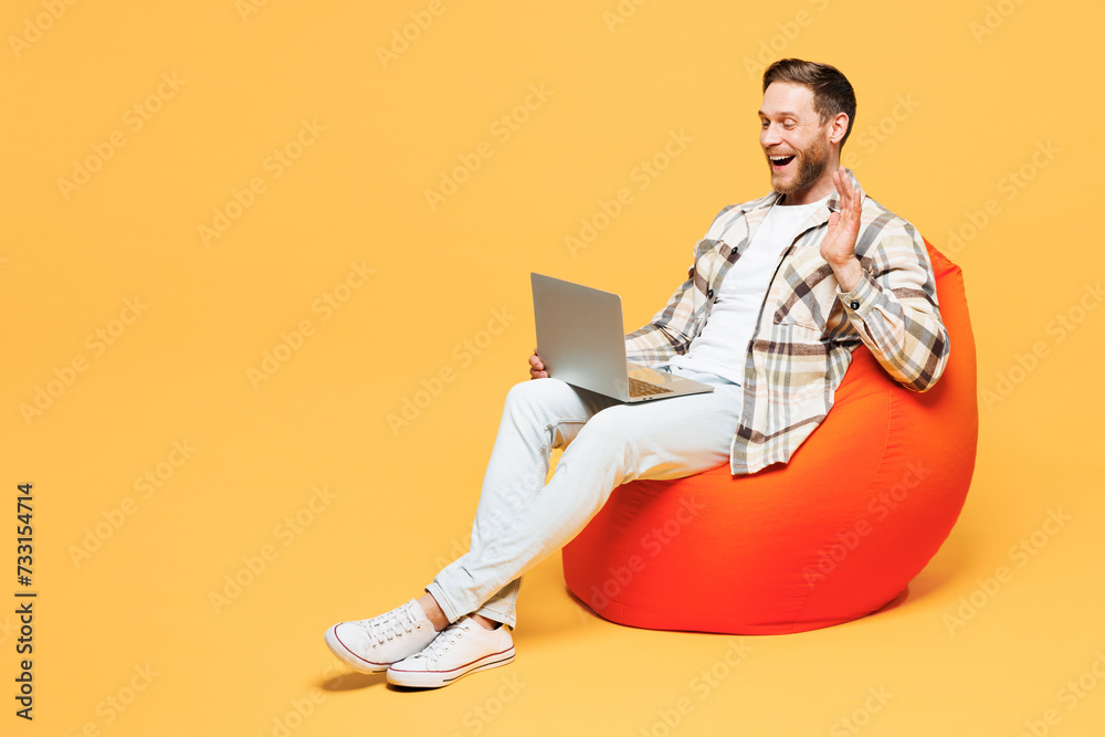 Full body young IT man he wear brown shirt casual clothes sit in bag chair hold use work on laptop pc computer waving hand isolated on plain yellow orange background studio portrait Lifestyle concept Full body young IT man he wear brown shirt casual clothes sit in bag chair hold use work on laptop pc computer waving hand isolated on plain yellow orange background studio portrait Lifestyle concept