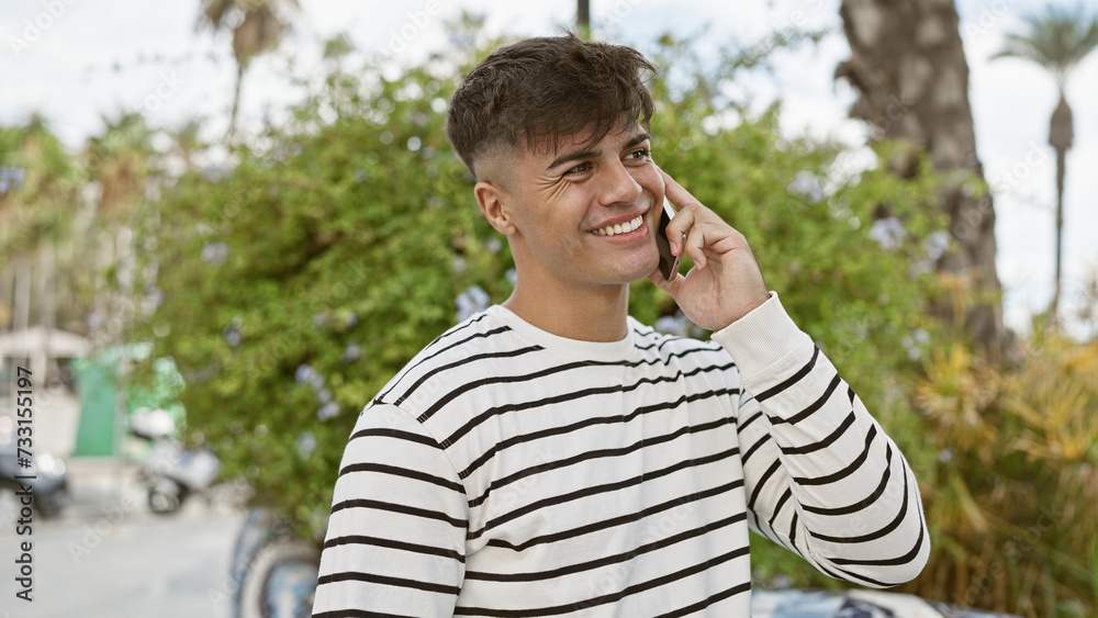 Confident young hispanic man, standing tall under the sunny park's green, smiling genuinely. with his casual fashion looking cool, talking on his smartphone, radiating happiness.