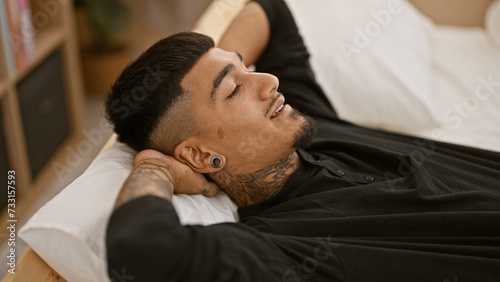 Handsome, tattooed young latin man lying on bed, hands resting on head, radiating confidence and relaxation as he rouses from sleep, a portrait of carefree indoor lifestyle