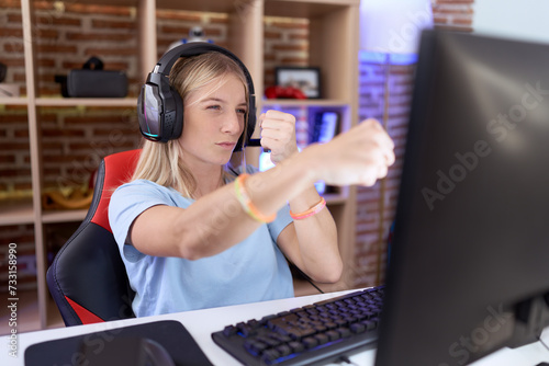 Young caucasian woman playing video games wearing headphones punching fist to fight, aggressive and angry attack, threat and violence