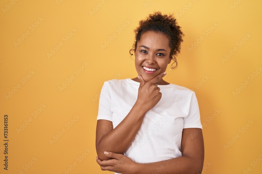 Young hispanic woman with curly hair standing over yellow background looking confident at the camera smiling with crossed arms and hand raised on chin. thinking positive. Young hispanic woman with curly hair standing over yellow background looking confident at the camera smiling with crossed arms and hand raised on chin. thinking positive.
