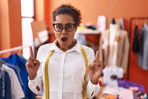 Young hispanic woman with curly hair standing at tailor room amazed and surprised looking up and pointing with fingers and raised arms.