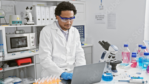 A young adult man with curly hair wearing lab coat and safety goggles works on a laptop in a modern laboratory setting.