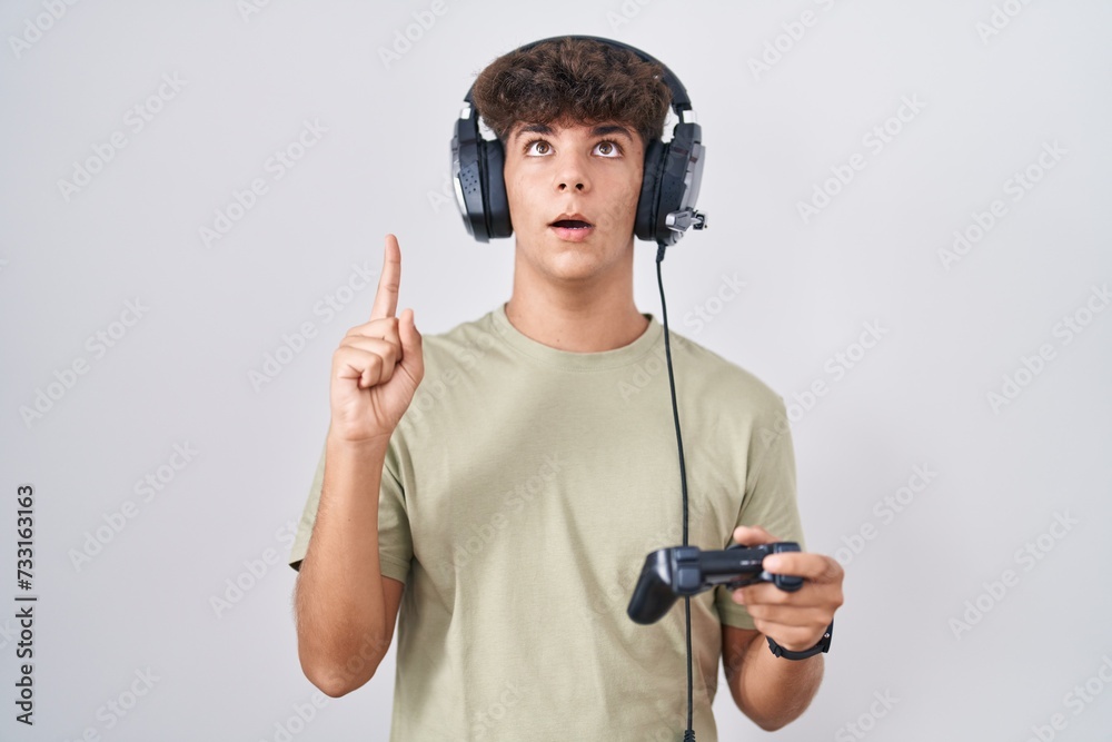 Hispanic teenager playing video game holding controller amazed and surprised looking up and pointing with fingers and raised arms. Hispanic teenager playing video game holding controller amazed and surprised looking up and pointing with fingers and raised arms.
