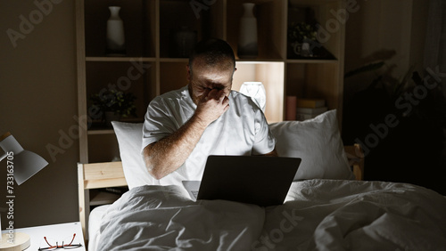 Exhausted caucasian man slumped over laptop while sitting on bedroom bed, stressed from overwork in dimly lit room