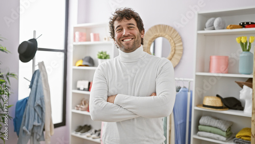 Obraz na plátne Handsome hispanic man with beard smiling in a modern wardrobe room, arms crossed, wearing a white sweater