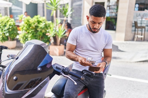 Young latin man counting dollars sitting on motorbike at street