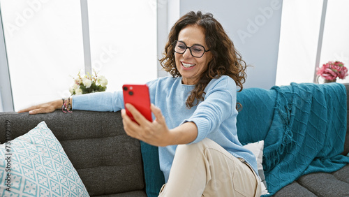 A smiling middle-aged hispanic woman with curly hair takes a selfie indoors on a couch, depicting a cozy home atmosphere. photo