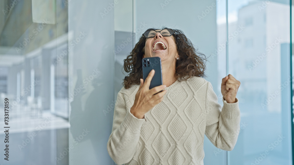 Fototapeta premium Excited hispanic woman celebrating with smartphone in modern office setting