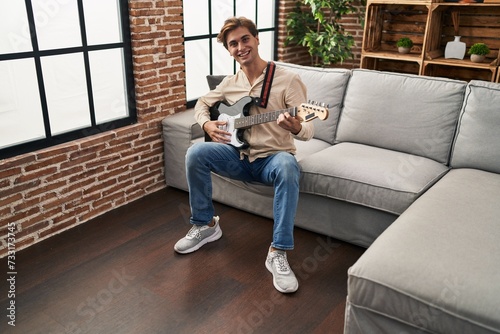 Young caucasian man playing electrical guitar sitting on sofa at home