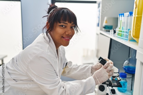 Young beautiful latin woman scientist smiling confident using microscope at laboratory