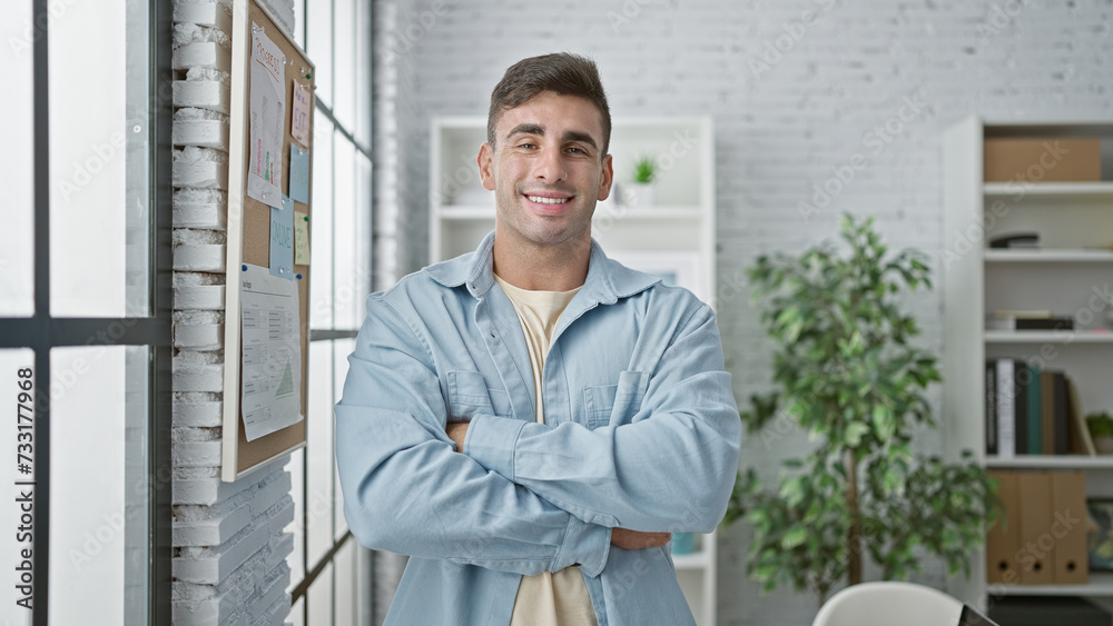 Joyful young hispanic man confidently standing with arms crossed at workplace, beaming as successful business worker in office interior, looking relaxed and positive at camera.