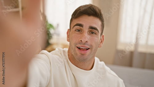 Cheerful young hispanic man sitting on bed, engaging in a heartwarming digital conversation via video call. captured in a cozy bedroom, showing the blend of modern technology, and home comfort.