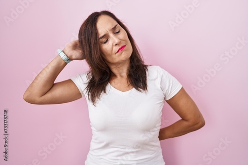 Middle age brunette woman standing over pink background suffering of neck ache injury, touching neck with hand, muscular pain