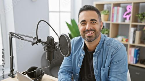 A smiling hispanic man with a beard in a studio setting with microphone and laptop, projecting a professional yet casual demeanor.