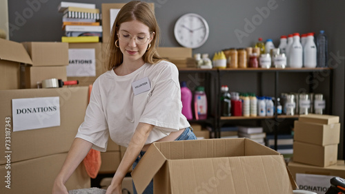 Young blonde woman volunteer packing clothes on cardboard smiling at charity center