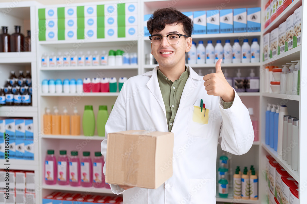 Young non binary man working at pharmacy drugstore holding box smiling happy and positive, thumb up doing excellent and approval sign Young non binary man working at pharmacy drugstore holding box smiling happy and positive, thumb up doing excellent and approval sign