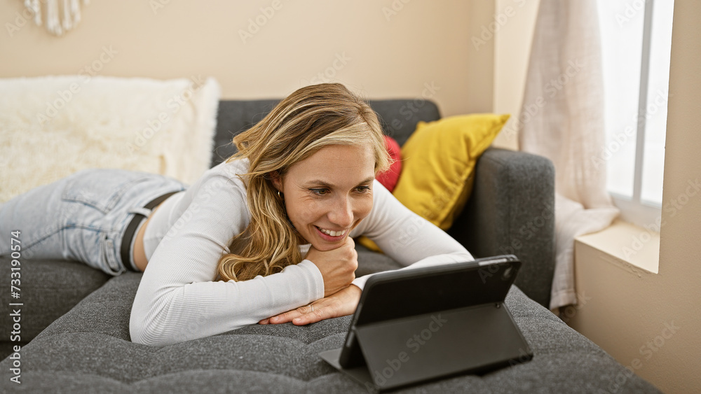 A smiling young woman lies on a couch using a tablet in a cozy, well-lit living room setting. A smiling young woman lies on a couch using a tablet in a cozy, well-lit living room setting.