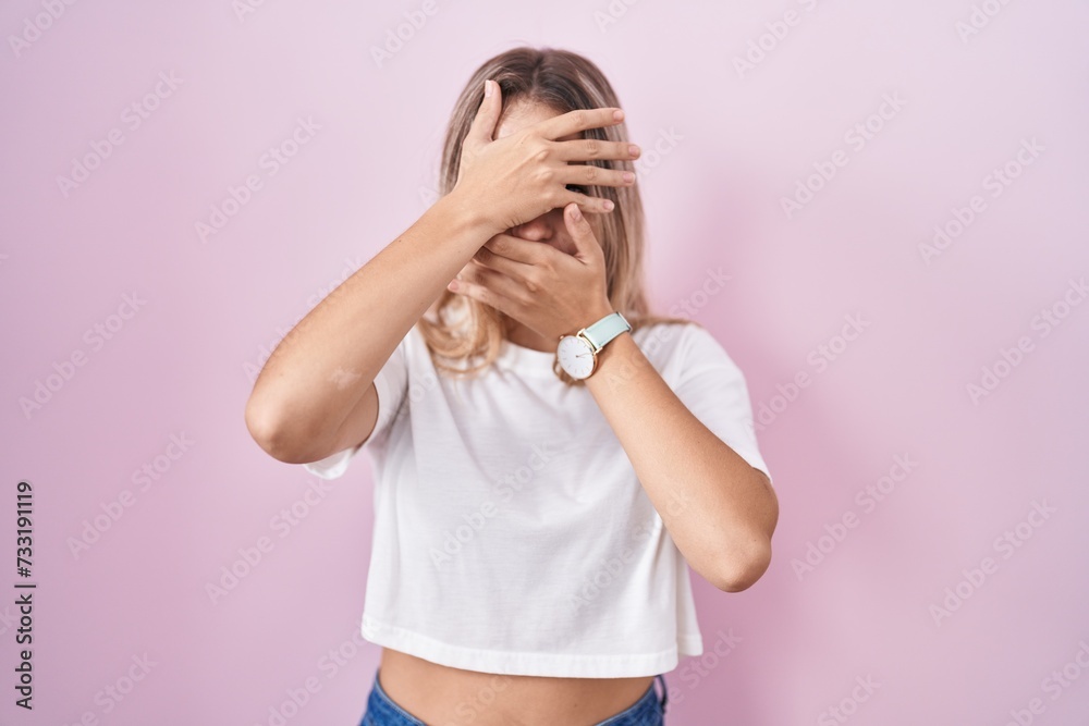 Young blonde woman standing over pink background covering eyes and mouth with hands, surprised and shocked. hiding emotion Young blonde woman standing over pink background covering eyes and mouth with hands, surprised and shocked. hiding emotion