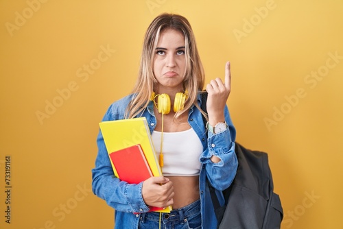 Young blonde woman wearing student backpack and holding books pointing up looking sad and upset, indicating direction with fingers, unhappy and depressed.