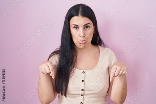 Young hispanic woman standing over pink background pointing down looking sad and upset, indicating direction with fingers, unhappy and depressed.