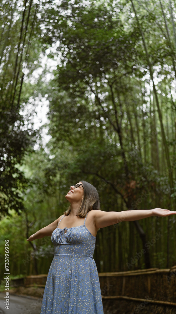 Naklejka premium Joyful hispanic woman embraces freedom in kyoto's bamboo forest, smiling, posing with open arms, looking around. beautiful, confident, carefree latin adult in glasses enjoying japan's green beauty.