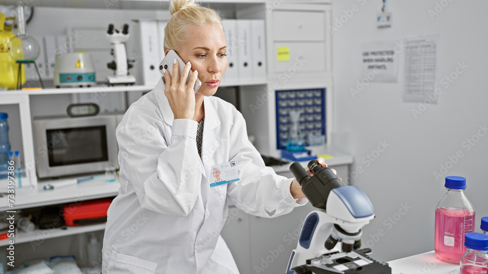 Intriguing talk, beautiful, serious young blonde scientist engages in tense conversation over smartphone while skillfully applying biology analysis using a microscope in laboratory