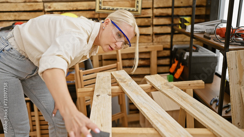 Young blonde woman carpenter wearing security glasses sanding wood plank at carpentry