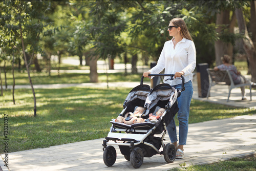 Lifestyle shot of young beautiful caucasian mother walking in green city park with two twin sisters in a double baby stroller. Happy Mothers day concept. Copy space.