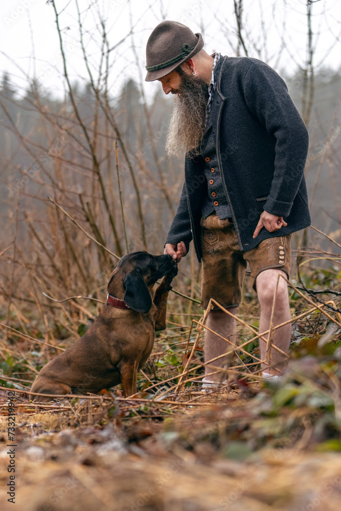 A bavarian man wearing a traditional folk costume interacting with his bavarian mountain dog nearby