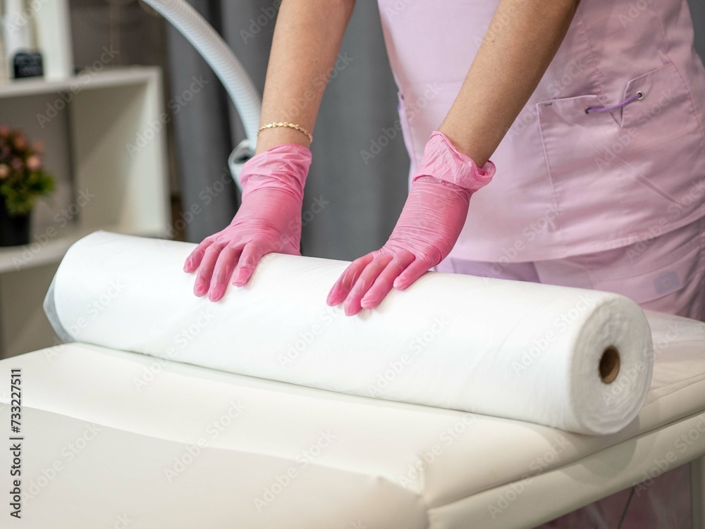 A master woman in a pink uniform in the cosmetology room spreads a white disposable spunbond sheet before the procedure. Preparing the cabinet for the client