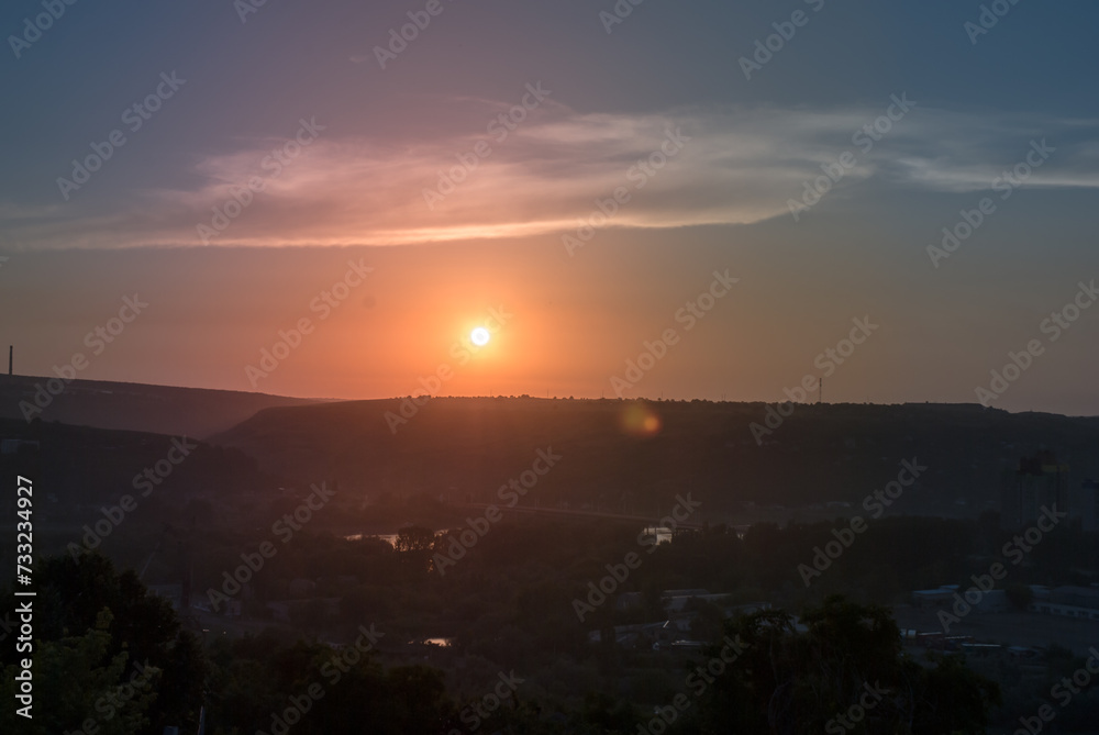A tranquil sunset over a valley with hints of industrial structures among greenery