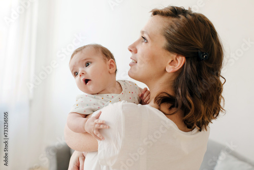 Side view of cheerful young brunette woman in casual clothes embracing precious infant child in sunlit living room and looking up photo