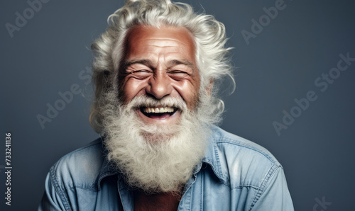 Radiant elderly man with flowing white beard and hair, wearing denim shirt, laughing heartily against a neutral grey background