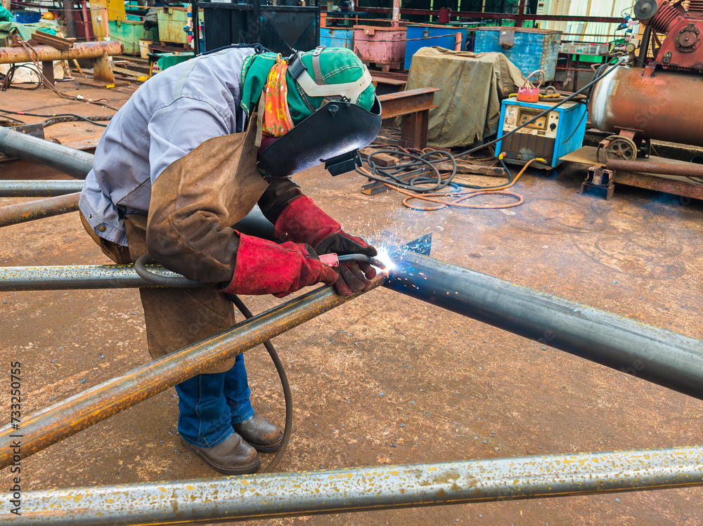 Welder Welding Steel Pipe for Steel Structure Work with Process Flux Cored Arc Welding (FCAW) and Appropriately Dressed with Personal Protective Equipment (PPE) for Safety, at an Industrial Factory. Welder Welding Steel Pipe for Steel Structure Work with Process Flux Cored Arc Welding (FCAW) and Appropriately Dressed with Personal Protective Equipment (PPE) for Safety, at an Industrial Factory.