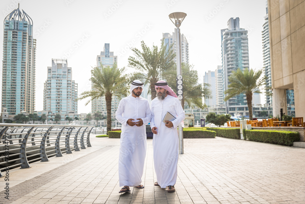 Arab businessmen wearing traditional emirati kandora meeting outdoors - Middle-eastern adults with dishdasha clothing bonding in Dubai