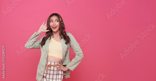 Young happy asian teen woman hand over ear listening an hearing to rumor or gossip and looking something away to side with smile on face isolated on pink copy space background.