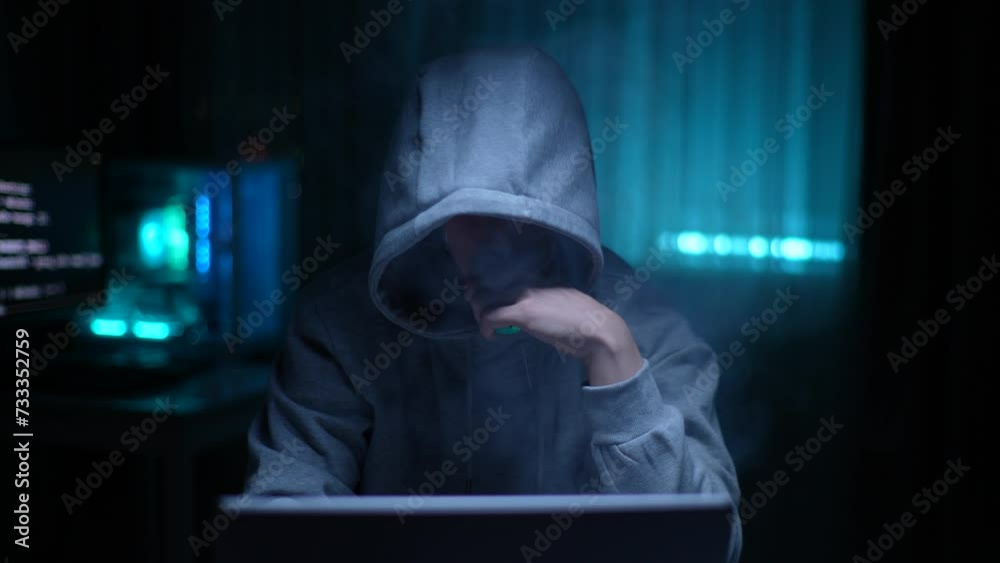Front view of unrecognizable male hacker in sweatshirt with hood vaping electronic cigarettes, writing code for cyber attack using using laptop computer, at desk in dark room with neon blue light Front view of unrecognizable male hacker in sweatshirt with hood vaping electronic cigarettes, writing code for cyber attack using using laptop computer, at desk in dark room with neon blue light