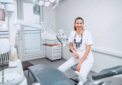 Portraait of sincerely smiling young dentist woman dressed white medical scrubs uniform sitting in modern dental clinic next to stomatology chair. Health care and medicare industry concept image.