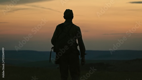 Silhouette of lonely soldier walking in field on red sunset background. A sad conceptual scene about the history of the soldiers of World War II and the Cold War.
