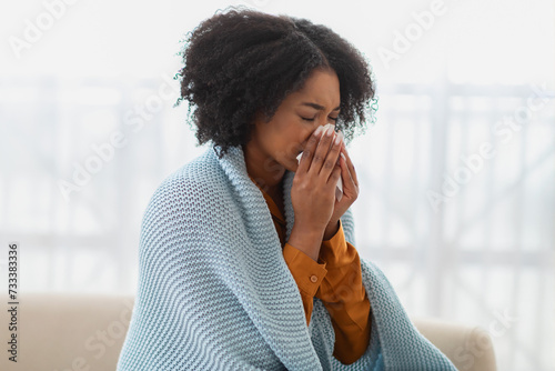 Sick woman with curly hair sneezing into a tissue, wrapped in a comforting blue blanket photo