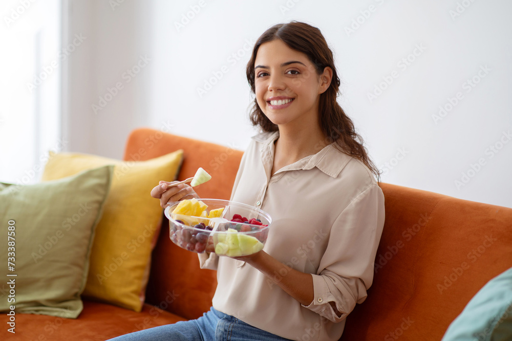 Cheerful woman holding bowl of fruit salad, enjoying healthy snack at home Cheerful woman holding bowl of fruit salad, enjoying healthy snack at home
