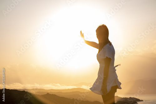 Young brunette girl in white dress touching the sun with one hand before it sets