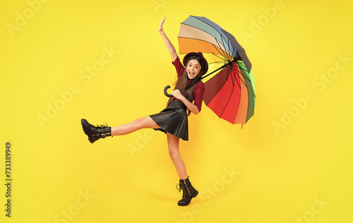 Excited teenager, glad amazed and overjoyed emotions. Teen girl under rainbow umbrella in autumn weather isolated on yellow background. Autumn kids clothes.