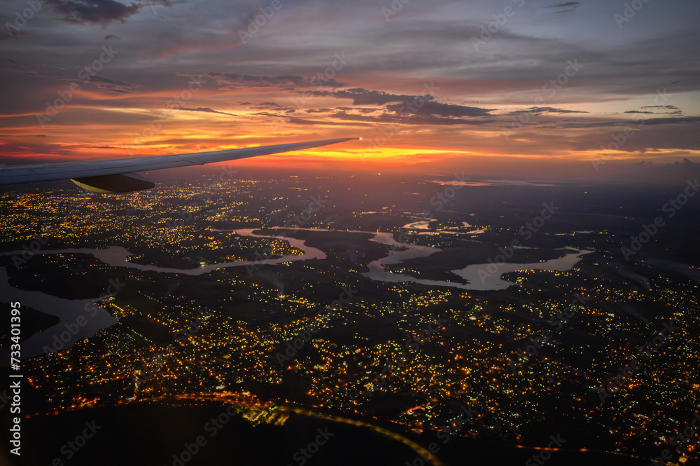 Vista da janela de avião comercial do Rio Iguaçu e Rio Paraná durante voo noturno com luzes da cidade e céu dramático em tons vermelho, roxo e azul. Foz do Iguaçu, Paraná. 2024.