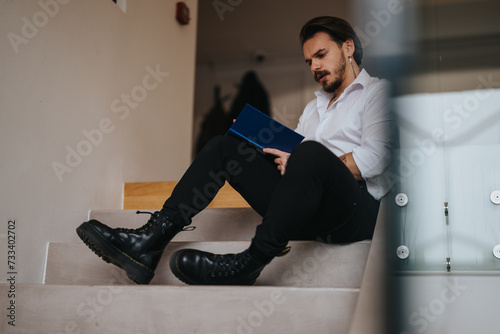 A young, professional man is engrossed in reading a blue folder while seated on a staircase, embodying focus and determination in a business setting.