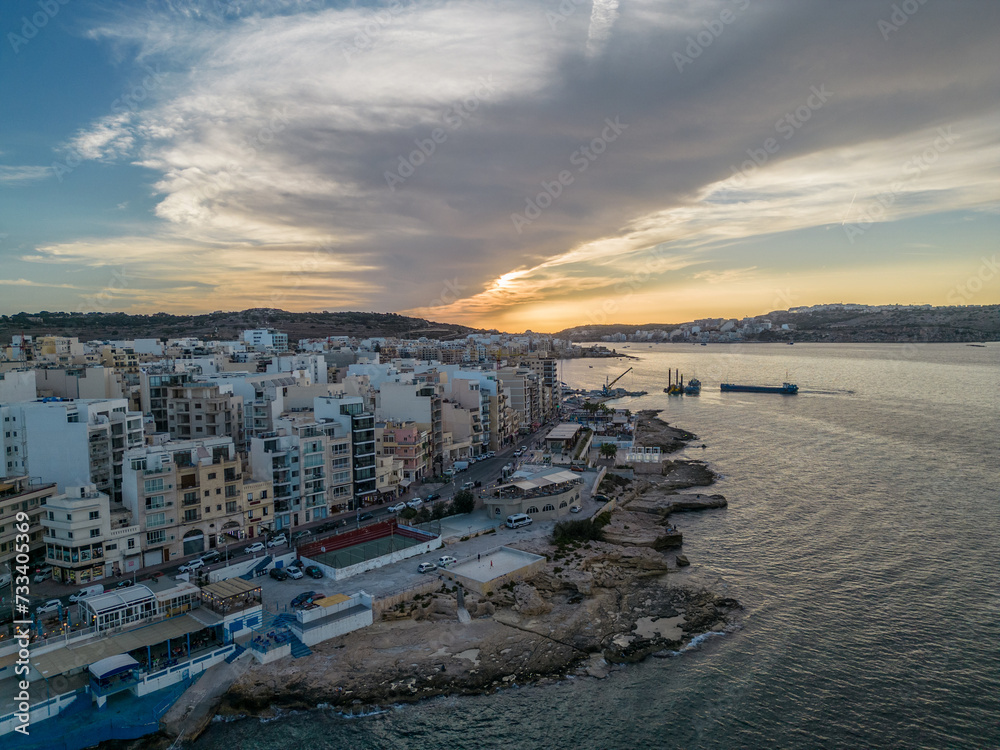 St. Paul's Bay at sunset with scenic cloud in Malta. Aerial view on the waterfront of Bugibba town and  rocky coastline at golden hour.